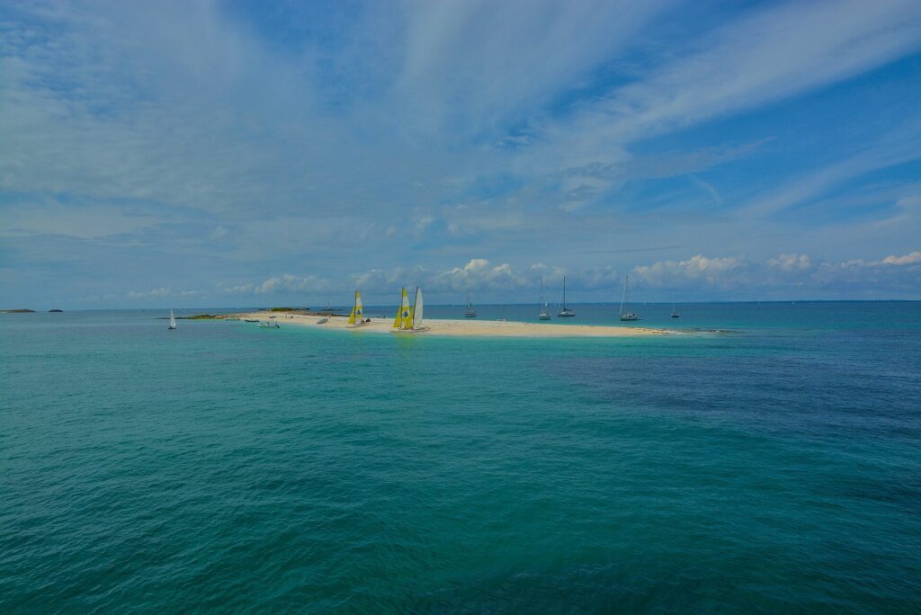 Een lange tong van wit zand met drooggevallen catamarans met gele en witte zeilen, omringd door zeilboten voor anker op turquoise water, onder een blauwe lucht met strepen wolken.