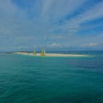 Een lange tong van wit zand met drooggevallen catamarans met gele en witte zeilen, omringd door zeilboten voor anker op turquoise water, onder een blauwe lucht met strepen wolken.