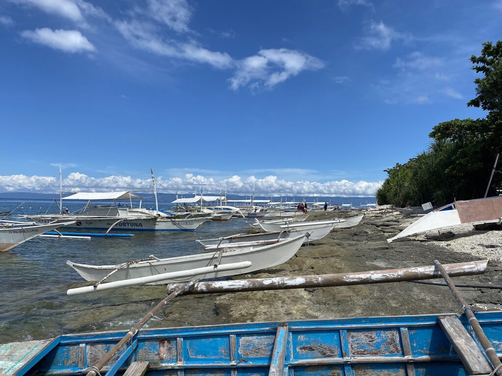 Een rij witte bangka's afgemeerd langs een rotsachtige kust, kleine prauwen met drijvers op de rotsen getrokken en een blauw bootje met afbladderende verf op de voorgrond, onder een blauwe lucht met witte wolken.