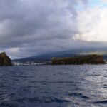 Vanaf een boot op een donkere, woelige zee gezien: een steil rotseilandje links en een lang, plat eilandje rechts, voor de stad Madalena en de hellingen van Pico onder een dreigende lucht