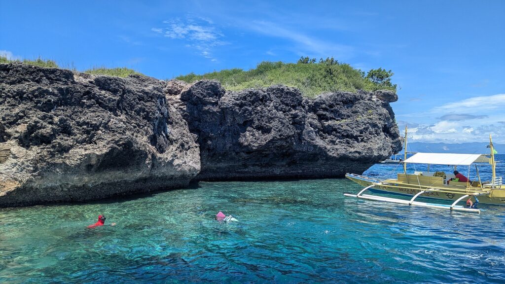 De grijze, uitgesleten kalksteenklif van het eiland Pescador, twee zwemmers met duikbril en snorkel en een gele banca in turquoise water