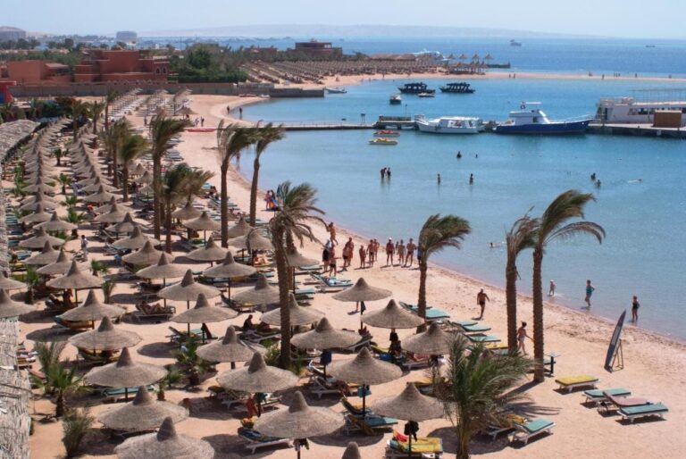 Het strand van het Giftun Azur Resort, met strooien parasols en palmbomen, en een steiger en boten op de achtergrond
