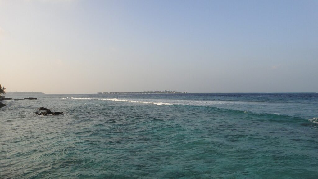 Vanaf een rotsachtige kust: een woelige turquoise pas waar een rij golven op het rif breekt, en aan de horizon een laag eiland met paalwoningen, onder een bleke lucht.