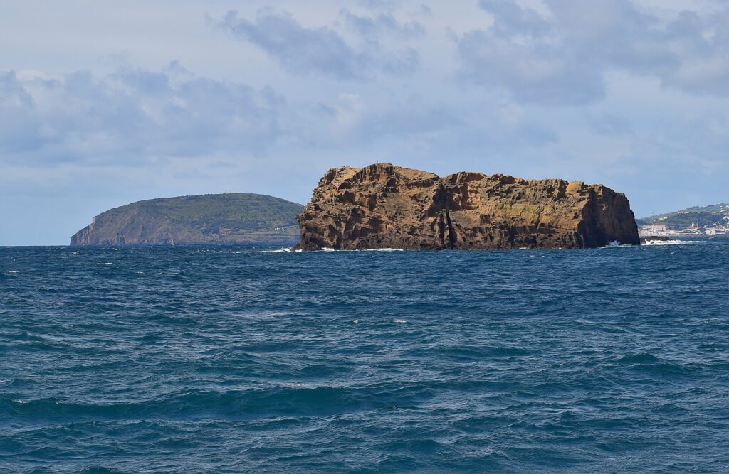 Het Ilhéu Deitado, een lang blok okerkleurig tuf met door holtes uitgesleten kliffen, op een woelige donkerblauwe zee, met erachter de groene heuvel van de Monte da Guia en verderop rechts huizen op Faial