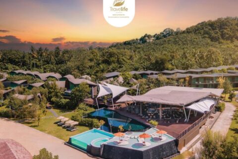 Luchtfoto van het Kalima Resort Khao Lak bij zonsondergang, met de zwembaden en de gebogen daken aan de voet van een beboste heuvel