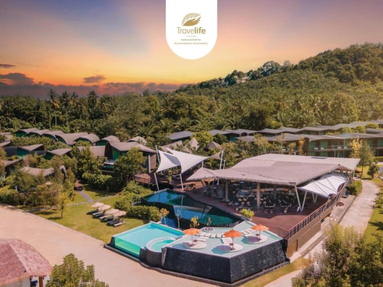 Luchtfoto van het Kalima Resort Khao Lak bij zonsondergang, met de zwembaden en de gebogen daken aan de voet van een beboste heuvel