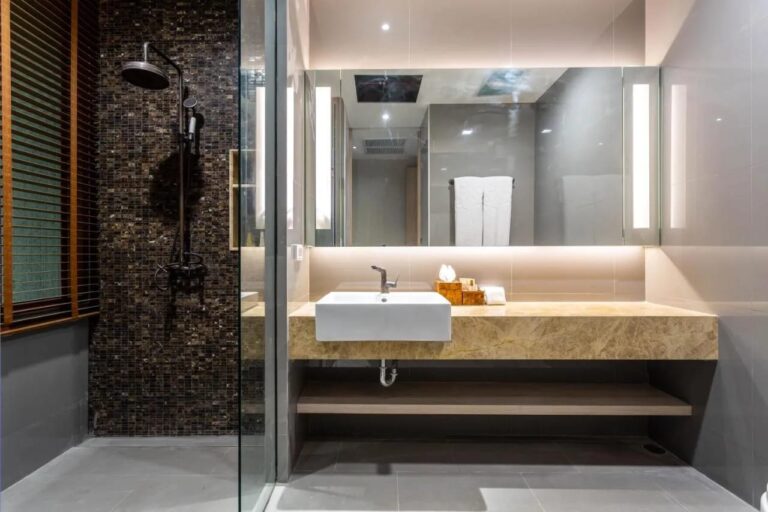 Een douche tegen een donkere mozaïekmuur, naast een waskom onder een verlichte spiegel