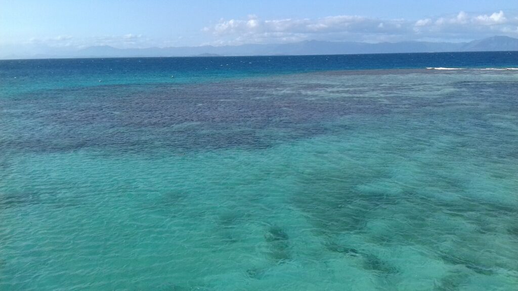 De lagune van bovenaf: turquoise water met donkere koraalblokken, rechts het barrièrerif waar de golven breken en aan de horizon de bergen van Grande Terre