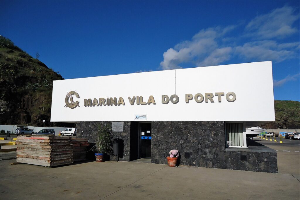 Het ontvangstgebouw van de jachthaven van Vila do Porto, een groot wit bord met metalen letters op een muur van zwart basalt, onder een blauwe lucht, links een groene klif