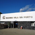 Het ontvangstgebouw van de jachthaven van Vila do Porto, een groot wit bord met metalen letters op een muur van zwart basalt, onder een blauwe lucht, links een groene klif