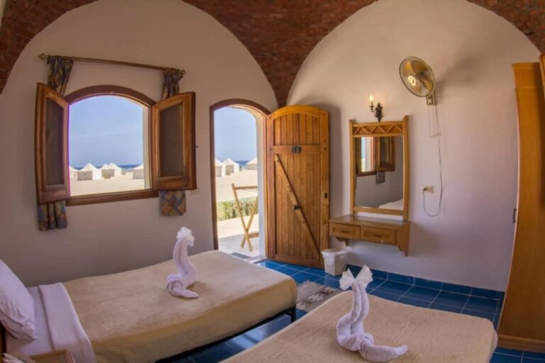 Een kamer met twee bedden die uitkomt op het strand, met witte tenten en de zee op de achtergrond