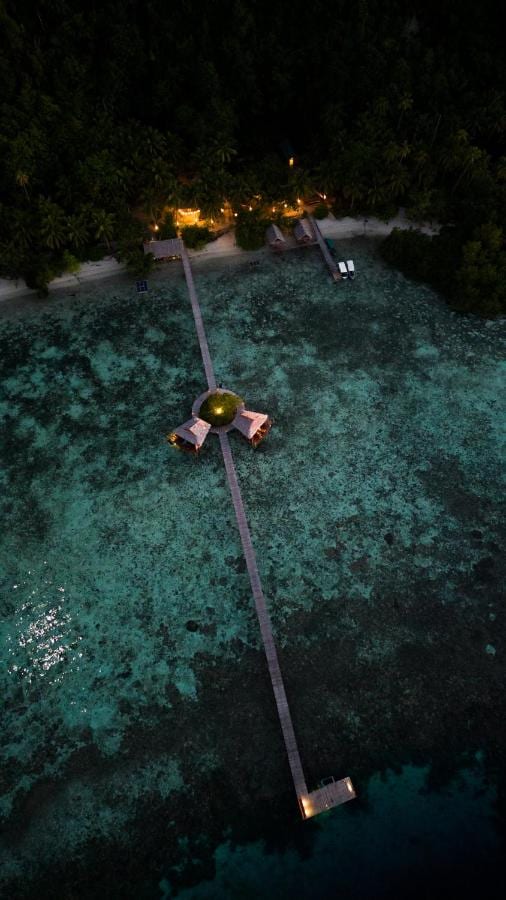 Luchtfoto bij nacht van een lange houten steiger vanaf het strand, met halverwege twee hutten op palen