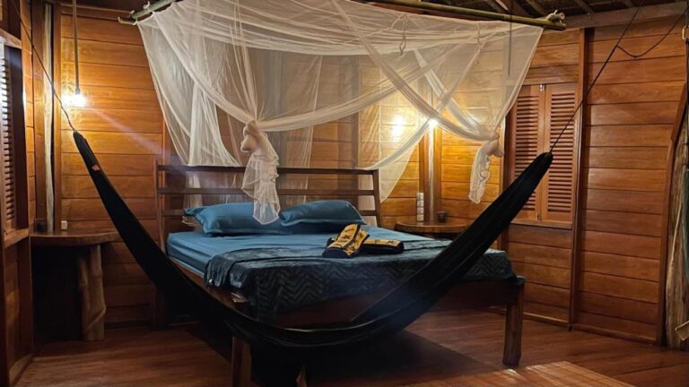 Een kamer met houten wanden, verlicht door lampen, met een bed onder een klamboe en een zwarte hangmat