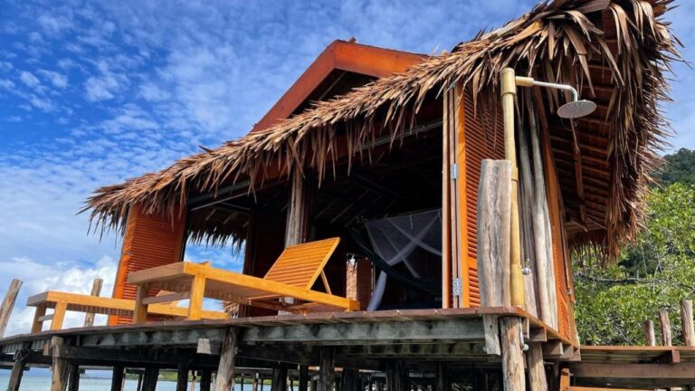 Een bungalow op palen met een dak van palmbladeren, twee houten ligstoelen en een buitendouche