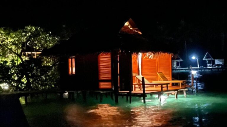 Een verlichte bungalow op palen bij nacht, met ligstoelen op het terras