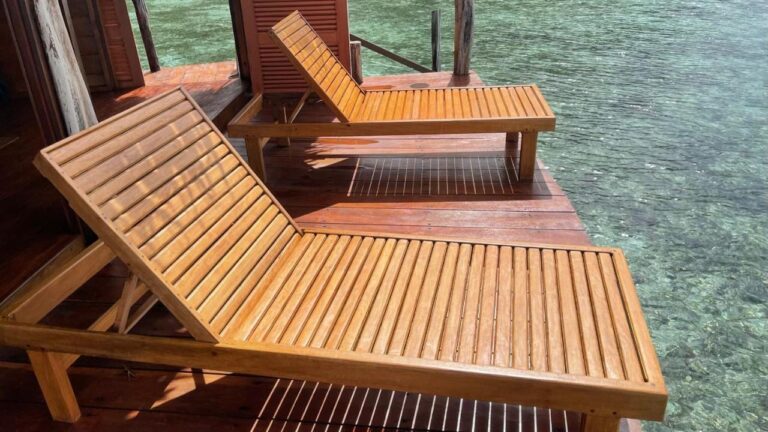 Twee houten ligstoelen op het terras van een bungalow, boven het heldere water