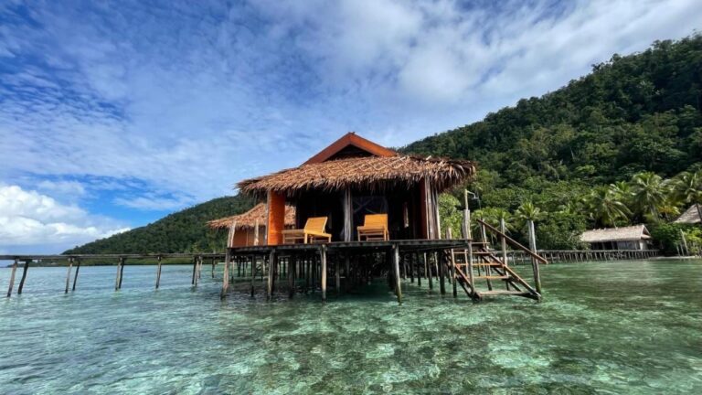 Een bungalow van Nyande Raja Ampat op palen boven het heldere water, aan de voet van een beboste heuvel
