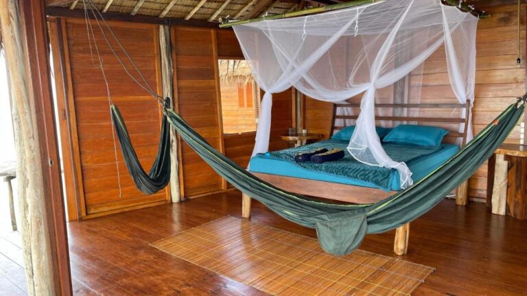Een houten kamer die openstaat naar buiten, met een bed met blauwe lakens onder een klamboe en een groene hangmat