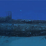 Onderwaterpanorama over de hele lengte van het wrak van de Rubis, rechtop op het zand, met de toren in het midden, in blauw water vol vissen.