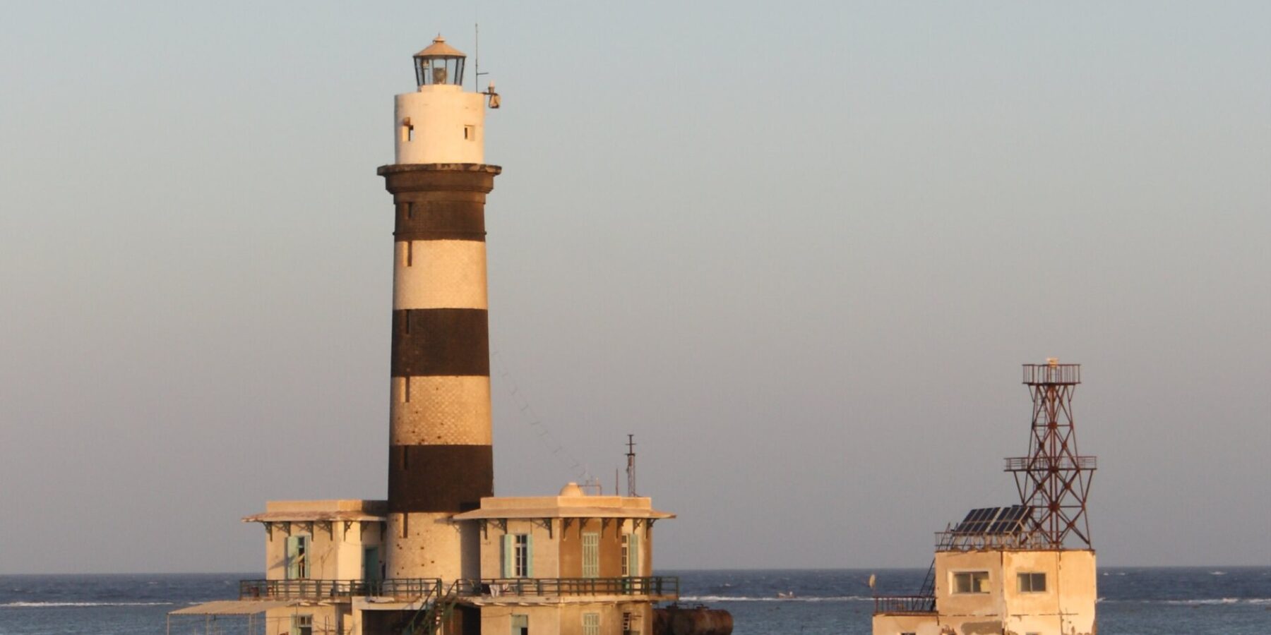 De vuurtoren van Daedalus, een zwart-wit gestreepte toren op het rif, met zijn gebouwen en metalen steiger, bij zonsondergang