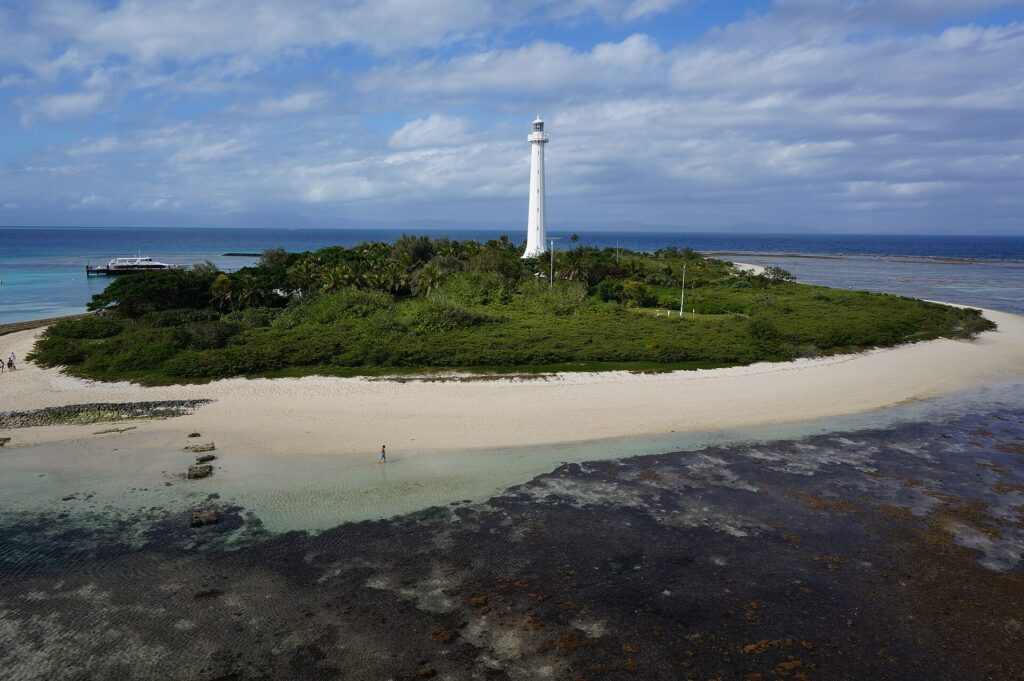 De grote witte vuurtoren van het eilandje Amédée rijst boven het groen uit, het eilandje omzoomd door wit zand, het drooggevallen koraalplateau op de voorgrond en links een aangemeerde boot