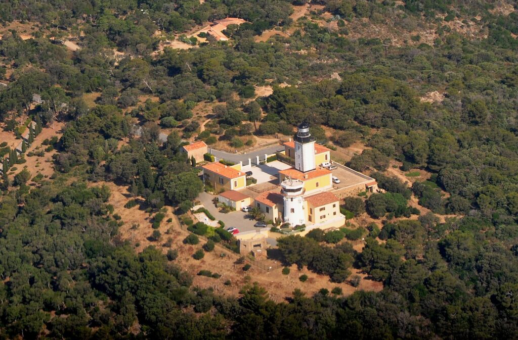 Luchtfoto van de vuurtoren van Cap Camarat, een witte toren met zwarte lantaarn, omringd door gele gebouwen met oranje pannendaken, midden in een dicht mediterraan bos.