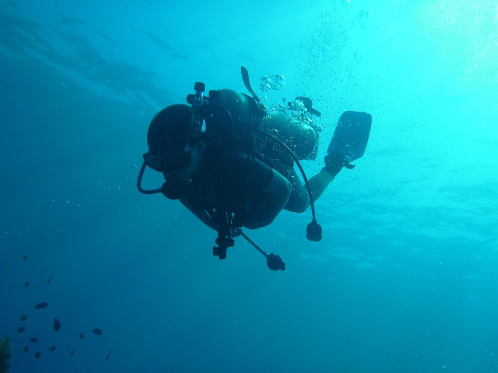Een duiker als silhouet van onderen gezien, fles, ademautomaten en vinnen afgetekend tegen het turquoise water, met zijn bellen die naar het verlichte oppervlak opstijgen.