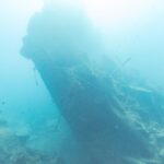 De gebroken achtersteven van de Thistlegorm steekt schuin omhoog in het blauw, met rechts een duiker