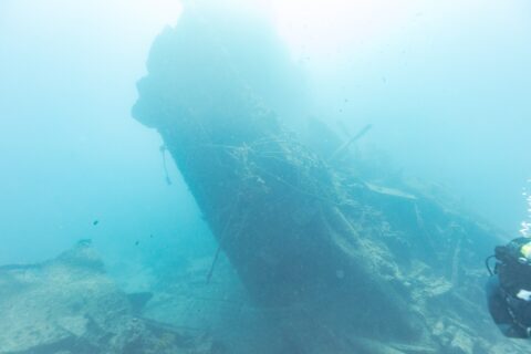 De gebroken achtersteven van de Thistlegorm steekt schuin omhoog in het blauw, met rechts een duiker