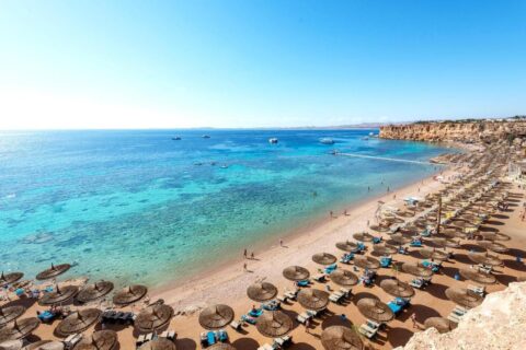 Een lang zandstrand aan de voet van een klif, met strooien parasols en een steiger in de turquoise zee