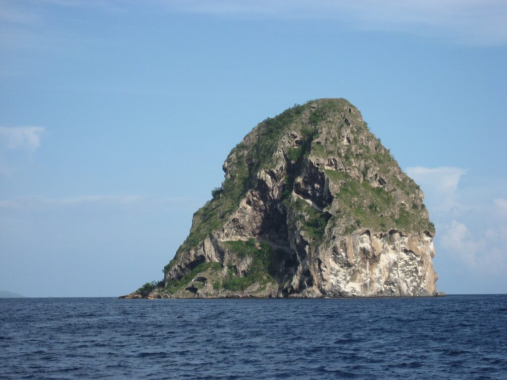 Diamond Rock van dichtbij gezien vanaf zee, een grijze rotsmassa met grotten, wit gestreept en begroeid op de top, boven een donkerblauwe zee