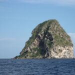 Diamond Rock van dichtbij gezien vanaf zee, een grijze rotsmassa met grotten, wit gestreept en begroeid op de top, boven een donkerblauwe zee