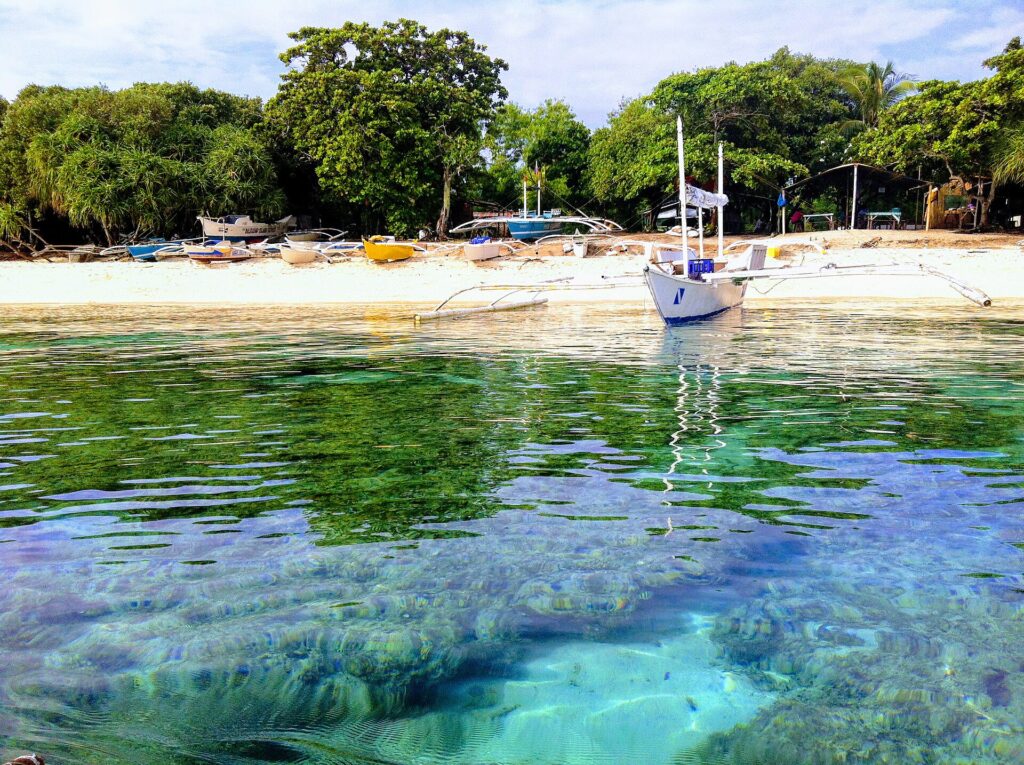 Vlak boven het water: koraalblokken onder helder turquoise water, voor een wit zandstrand waar een witte bangka voor anker ligt, andere gekleurde bootjes onder de bomen op het strand getrokken.