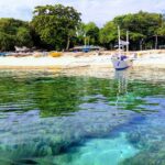 Vlak boven het water: koraalblokken onder helder turquoise water, voor een wit zandstrand waar een witte bangka voor anker ligt, andere gekleurde bootjes onder de bomen op het strand getrokken.