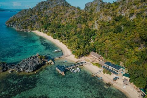 Luchtfoto van het Sangat Island Dive Resort, met zijn strand, steiger en boten, aan de voet van de kliffen