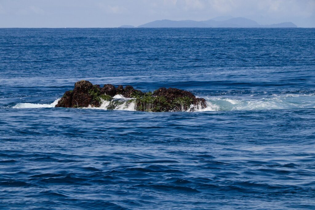De top van Richelieu Rock steekt bij laagwater boven het water uit, een donkere rots met groene algen waar de golven op slaan, met de Surin-eilanden aan de horizon