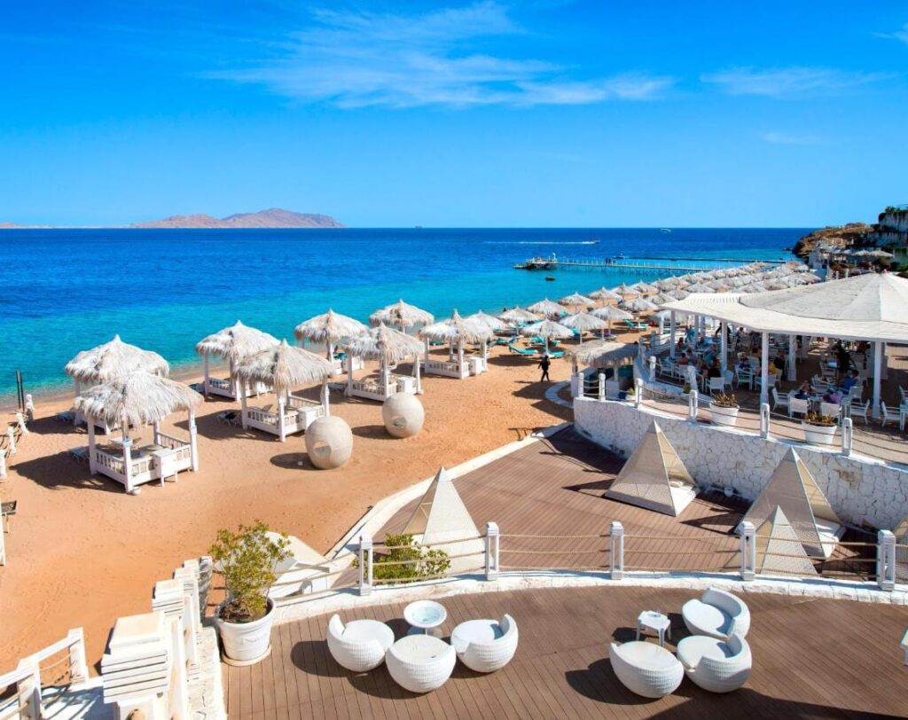 Het strand van het Sunrise Arabian Beach Resort, met strooien parasols en een steiger, tegenover het eiland Tiran