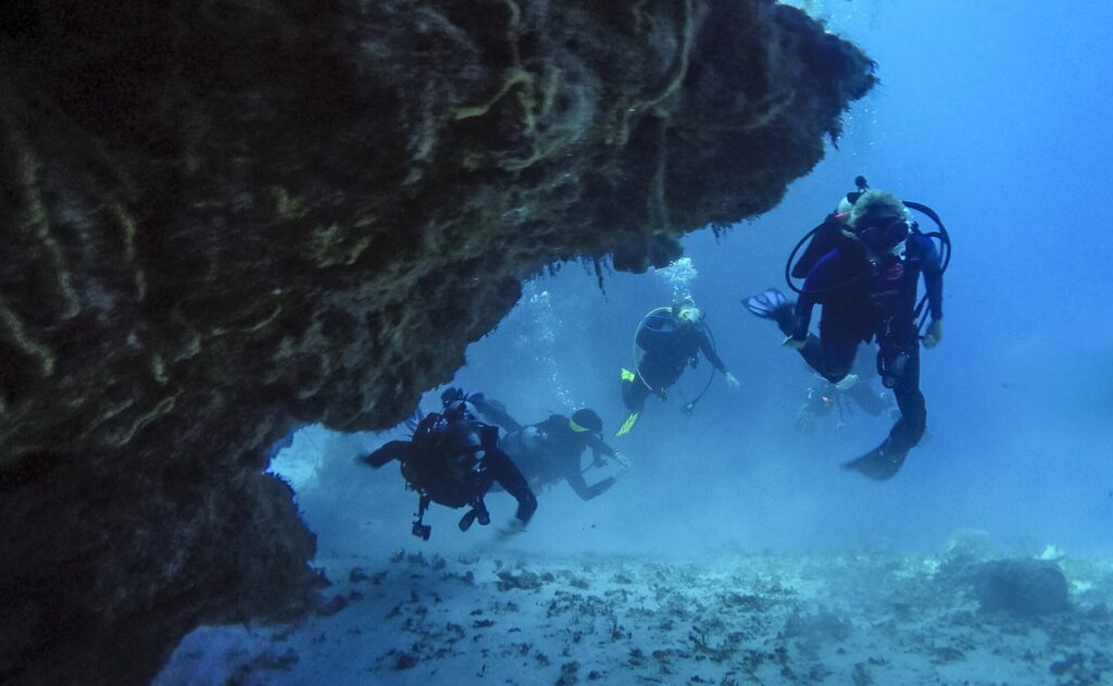 Vier duikers komen uit een koraaltunnel boven een zandbodem, bij Cozumel, met het rotsplafond boven hen