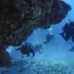 Vier duikers komen uit een koraaltunnel boven een zandbodem, bij Cozumel, met het rotsplafond boven hen