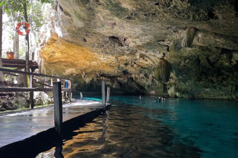 Het oostelijke oog van cenote Dos Ojos: een kalkstenen gewelf vol stalactieten boven turquoise water, links een houten steiger en achterin zwemmers