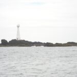 Een slanke witte vuurtoren met rode deur staat op het lage, rotsige eilandje Formigão, met donkere rotsen omzoomd door bruine algen, boven een grijze, woelige zee onder een bewolkte lucht