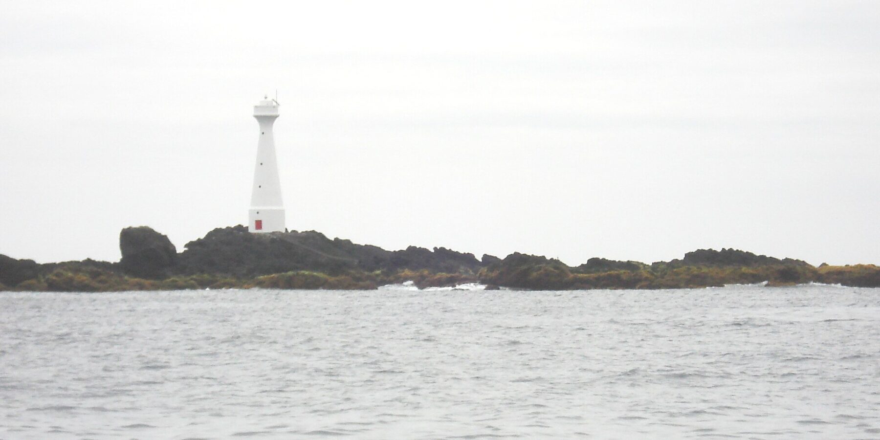 Een slanke witte vuurtoren met rode deur staat op het lage, rotsige eilandje Formigão, met donkere rotsen omzoomd door bruine algen, boven een grijze, woelige zee onder een bewolkte lucht
