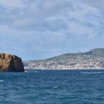 Panorama van de twee okerkleurige tufeilandjes van Madalena op een donkerblauwe zee, het lange, platte Ilhéu Deitado links en het rechtopstaande Ilhéu em Pé rechts, met de witte stad Horta en de groene heuvels van Faial op de achtergrond