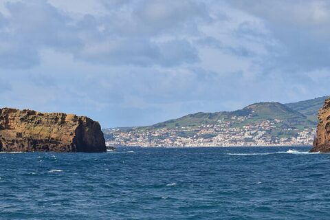 Panorama van de twee okerkleurige tufeilandjes van Madalena op een donkerblauwe zee, het lange, platte Ilhéu Deitado links en het rechtopstaande Ilhéu em Pé rechts, met de witte stad Horta en de groene heuvels van Faial op de achtergrond