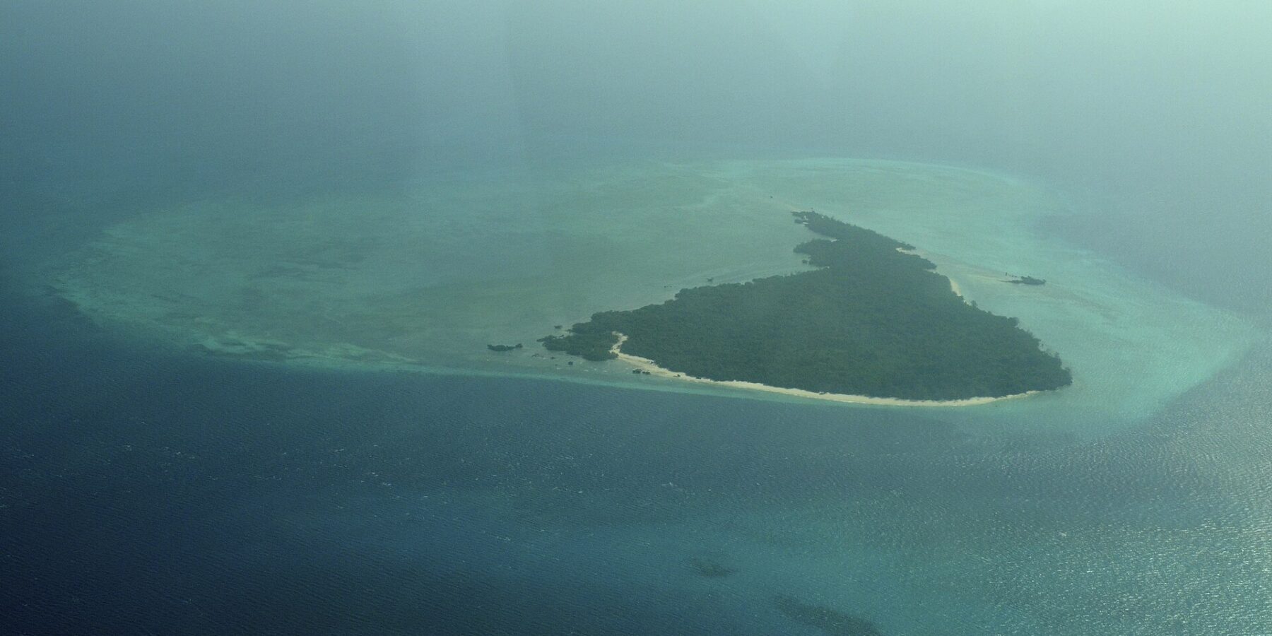 Het eiland Misali vanuit een vliegtuig, een driehoek van bos met een wit strand eromheen, op een turquoise rifplateau midden in een donkerblauwe zee, in de nevel