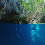 Half boven, half onder water bij de ingang van cenote The Pit: bovenaan het kalkstenen gewelf en de houten trap onder de jungle, onderaan twee duikers in het diepblauwe water