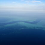 Luchtfoto van een donkerblauwe S-vormige geul die tussen de turquoise rifplateaus van het barrièrerif slingert, met een paar kleine witte boten in de pas