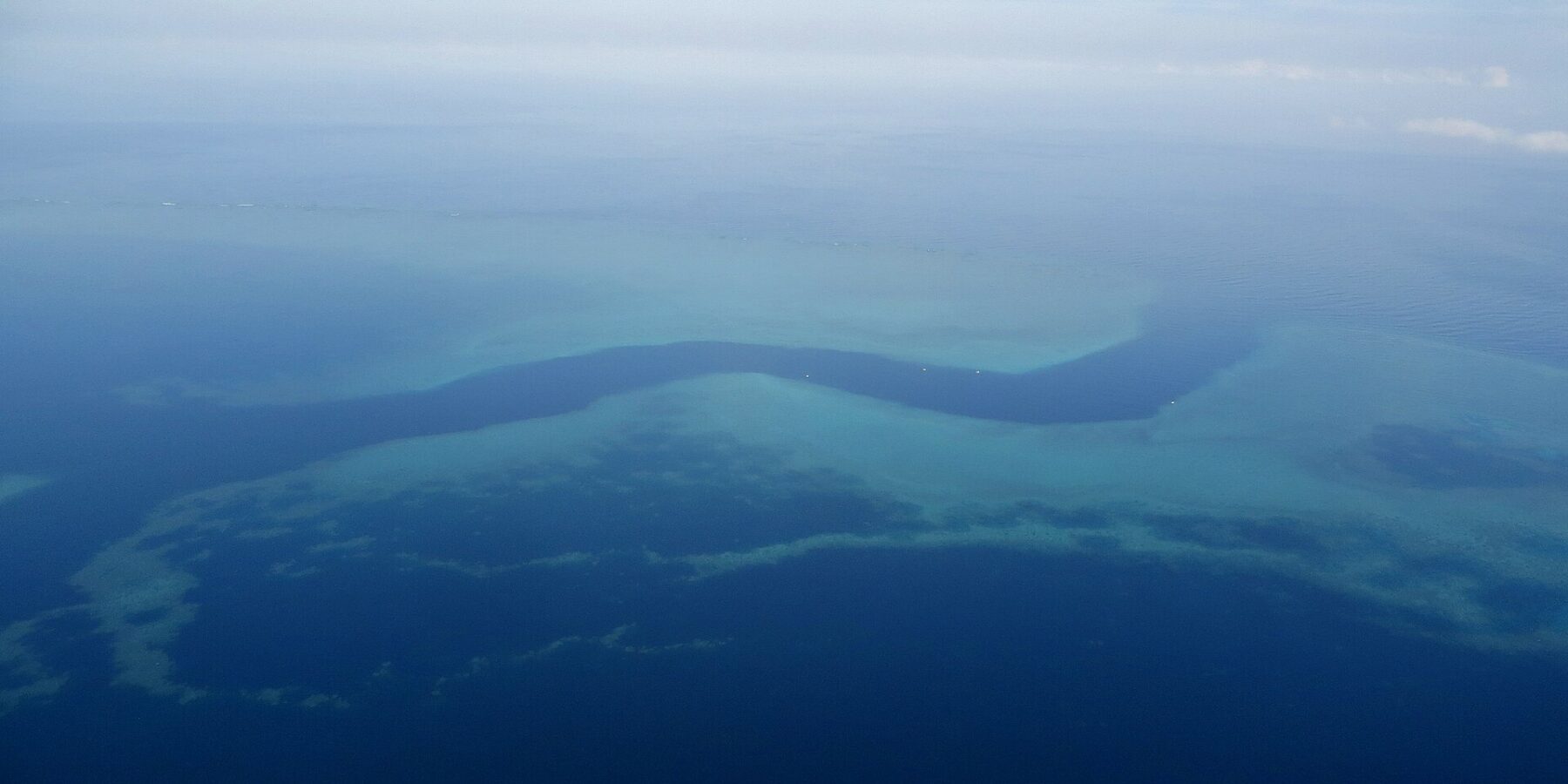 Luchtfoto van een donkerblauwe S-vormige geul die tussen de turquoise rifplateaus van het barrièrerif slingert, met een paar kleine witte boten in de pas