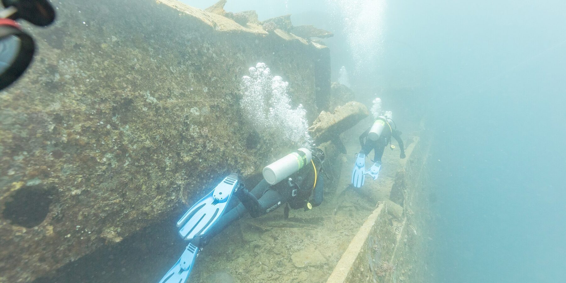 Twee duikers zwemmen over het dek van de Thistlegorm langs een gang, in melkachtig blauw water