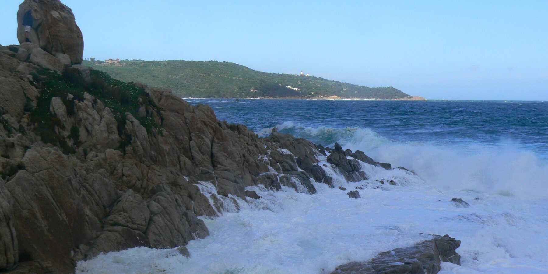 Golven breken in wit schuim op de granietrotsen van L'Escalet op de voorgrond, tegenover een woelige donkerblauwe zee, met in de verte de beboste kust van Cap Camarat en zijn witte vuurtoren.
